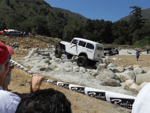 1986 Ford Bronco - An off-road event features a white wagon tackling a challenging rocky obstacle course under a clear blue sky.