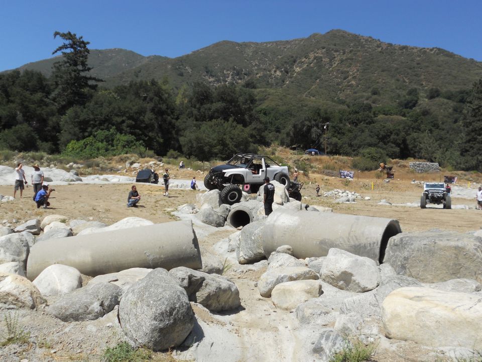 Modified off-road truck with large tires on rocks, other vehicles and people in background
