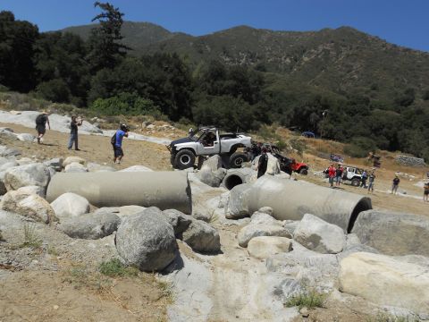 1986 Ford Bronco - Multiple off-road vehicles are seen navigating a challenging course with large concrete pipes and rocks.