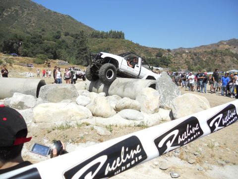 1986 Ford Bronco - A white off-road truck maneuvers over large concrete pipes during an event, with a crowd gathered to watch.