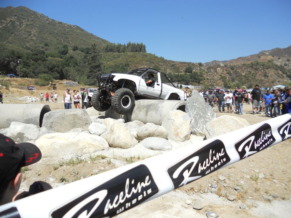 White off-road truck with large tires climbing over concrete pipes and rocks, with spectators watching