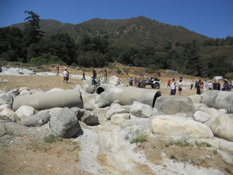 1986 Ford Bronco - People watch as an off-road vehicle navigates a challenging course with large rocks and concrete pipes.