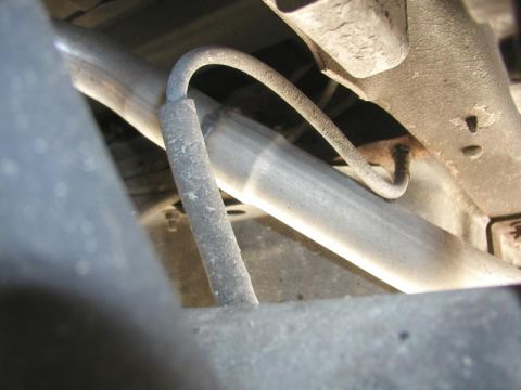 1988 Ford Bronco - Close-up under a vehicle showing a hard brake line looping down from a chassis bracket and running alongside a rusted axle housing or frame…