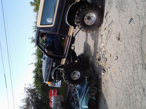1986 Ford Bronco - A lifted 1986 Ford Bronco is shown crushing a car, with the driver visible behind the wheel.
