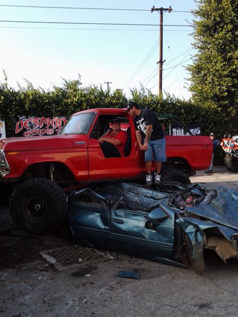 1986 Ford Bronco - A lifted red truck is parked on top of a severely damaged blue sedan.