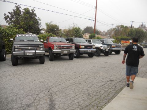 1986 Ford Bronco - A group of modified trucks and SUVs are lined up in a parking lot under an overcast sky.