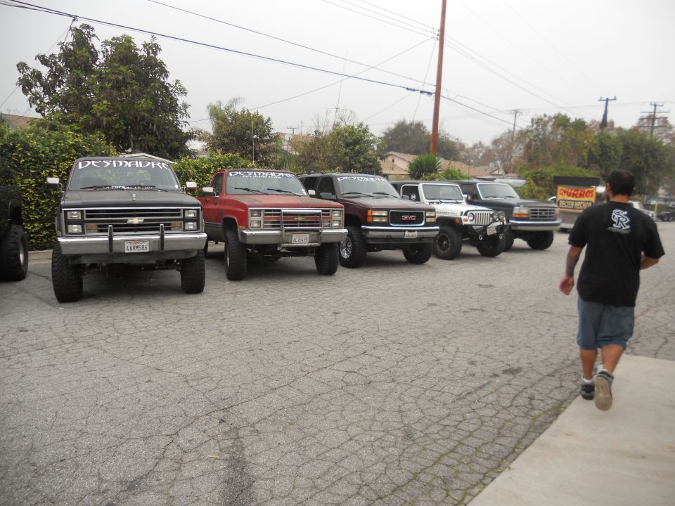 Several lifted trucks and SUVs parked in a lot, including a black Chevy, a red Chevy, a maroon GMC, a white Jeep, and a black Ford. A person walks away from the camera