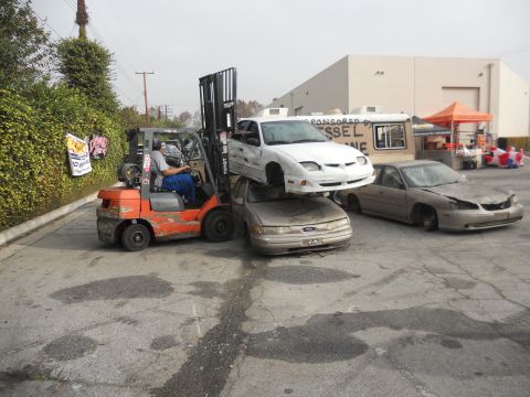 1986 Ford Bronco - A forklift stacks a white car on top of a tan Ford Taurus, with another car to the right.