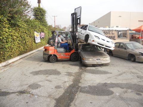 1986 Ford Bronco - A forklift is shown stacking a white car on top of a tan sedan, with another car visible in the background.