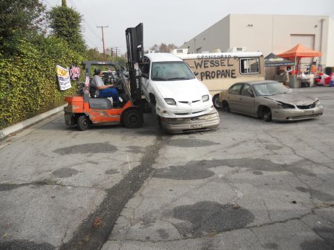 1986 Ford Bronco - A forklift is lifting a white car that has sustained significant front-end damage. Another damaged car is parked next to it.