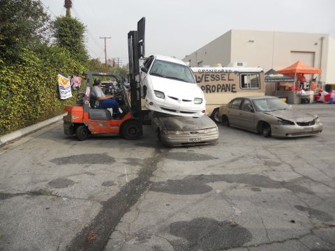 1986 Ford Bronco - A forklift is stacking cars in a lot, with a propane truck in the background.
