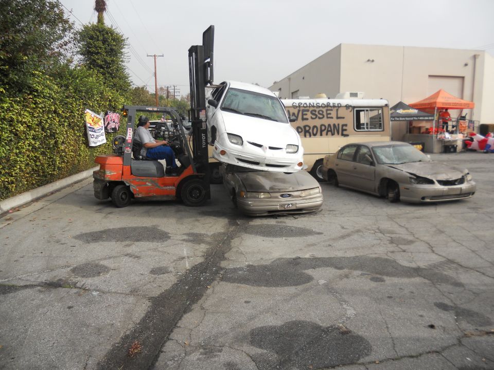 forklift lifting a white car onto a gold car, another gold car sits nearby