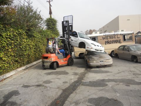 1986 Ford Bronco - A forklift is used to stack a white car on top of a tan car, with a propane truck in the background.