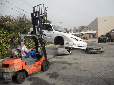 1986 Ford Bronco - A forklift is lifting a white car, which is suspended over two other vehicles in a yard.