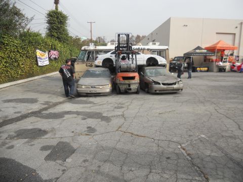 1986 Ford Bronco - A gold sedan with a damaged front end is parked on cracked asphalt, with other vehicles and a forklift in the background.
