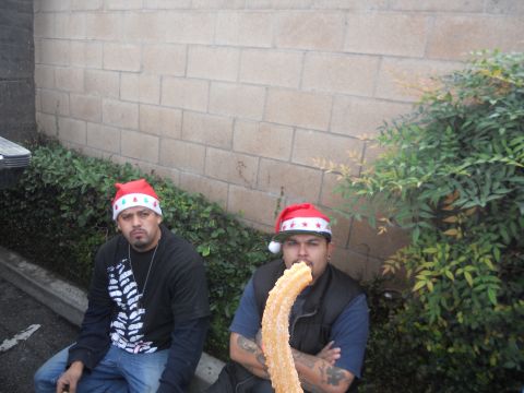 1986 Ford Bronco - Two men in Santa hats sit near a brick wall, one holding a large churro, with a "SUPERMOTORS" watermark in the corner.