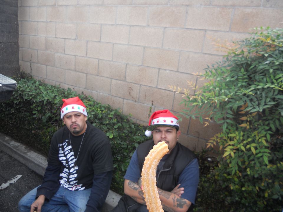 Two men in Santa hats sit near a brick wall, one holding a large churro