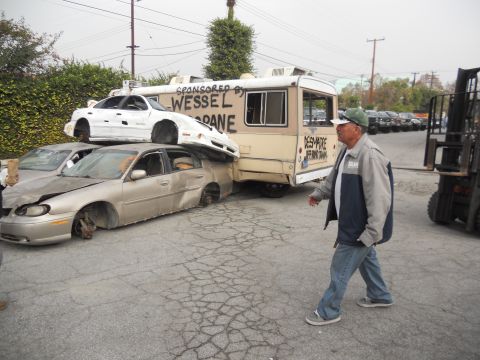 1986 Ford Bronco - Multiple damaged vehicles are piled up and leaning against a trailer in a lot.