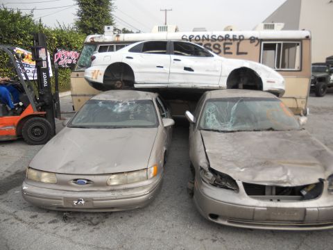 1986 Ford Bronco - A damaged gold Ford Taurus sits in a junkyard next to other vehicles.