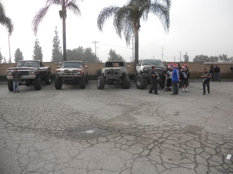 1986 Ford Bronco - Several modified trucks are parked in a lot, some with people gathered around.