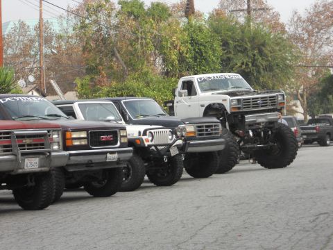 1986 Ford Bronco - Several modified trucks and SUVs are parked in a line, with a heavily lifted white Ford F-Series truck prominently displayed.