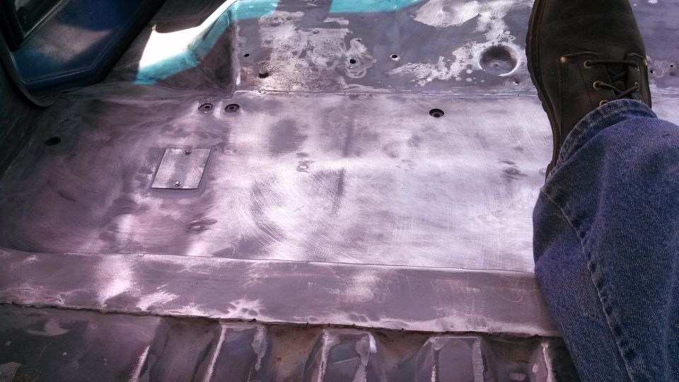 Bare metal floor pan of a vehicle interior, showing some rust and repair patches. A person's leg and boot are visible on the right