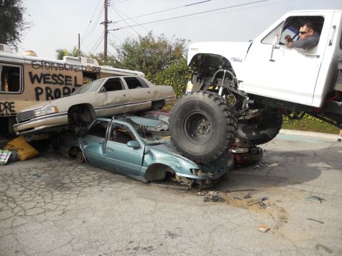 1986 Ford Bronco - A monster truck is crushing several cars, with a graffiti-covered trailer in the background.