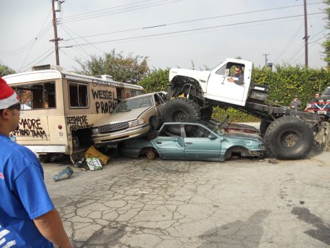 1986 Ford Bronco - A monster truck is piled on top of several wrecked cars, with a bus visible behind them.