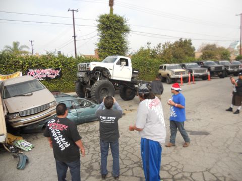 1986 Ford Bronco - A heavily modified white truck looms over damaged cars, with onlookers and other vehicles present at an outdoor event.