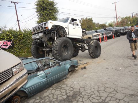 1986 Ford Bronco - A lifted monster truck is shown crushing a sedan, with other vehicles lined up in the background.
