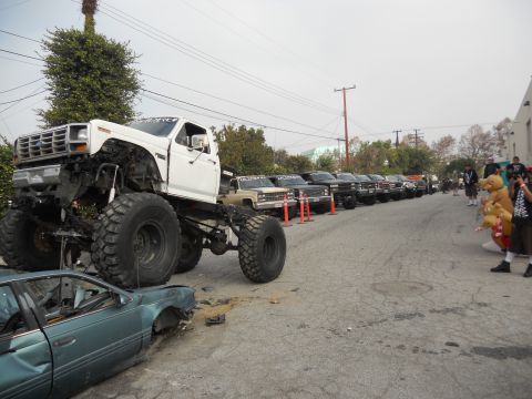 1986 Ford Bronco - Monster truck dominates the scene, crushing a sedan while other trucks line the street.