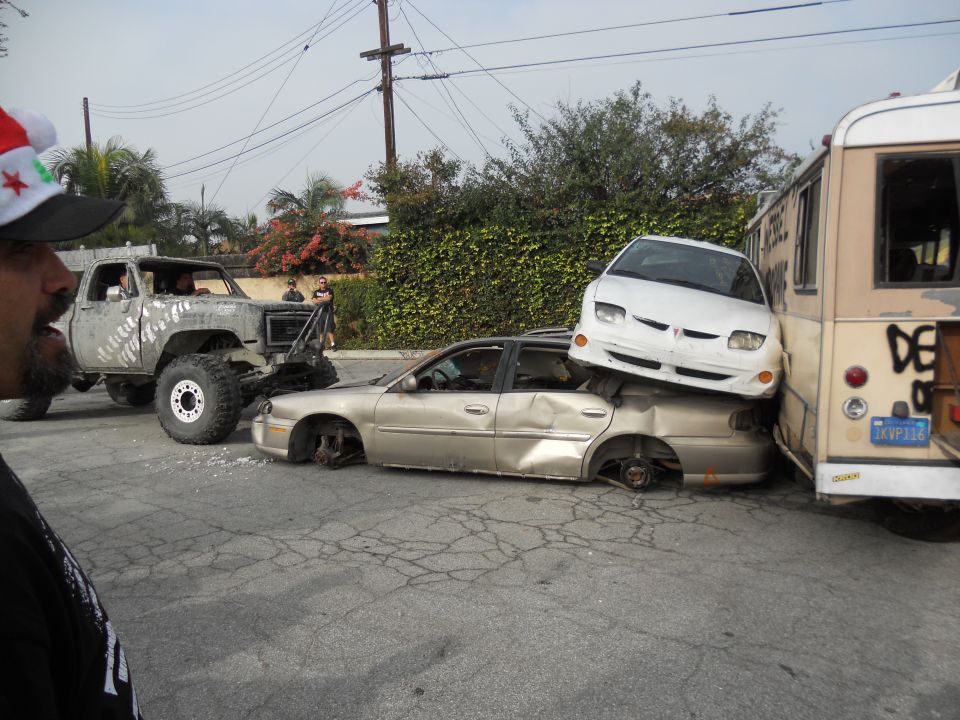 lifted pickup truck, a crushed sedan, a white car on top of the sedan, and a bus are in a junkyard scene