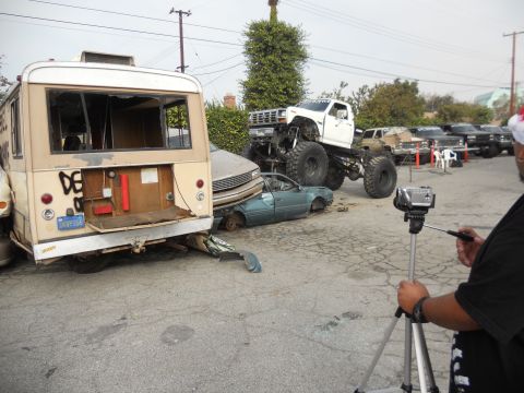 1986 Ford Bronco - A monster truck is crushing a car while a bus sits nearby, with a person filming the event.