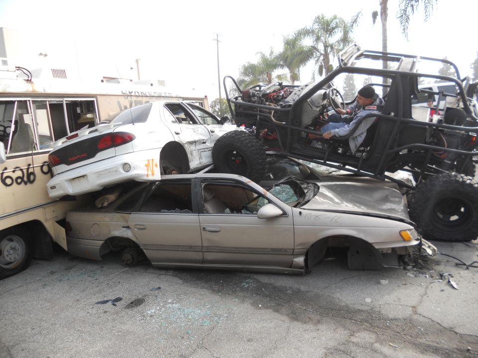 Several wrecked cars are piled up, with a custom off-road vehicle on top. A person is in the driver's seat of the off-road vehicle