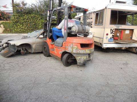 1986 Ford Bronco - Forklift moves a wrecked car near a bus with visible text. The car's front end is heavily damaged.