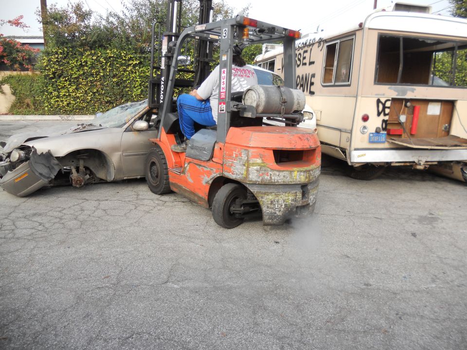 Damaged sedan being lifted by an orange Toyota forklift, with a beige bus in the background. Text on bus reads 'DIESEL PANE'