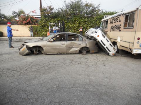 1986 Ford Bronco - A sedan is severely damaged, crushed by a truck, with a forklift and people present at the scene.