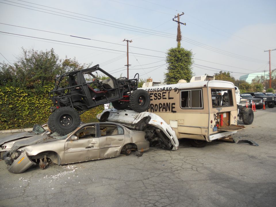 Off-road buggy on top of crushed cars next to a bus with 'VESSEL PROPANE' written on it