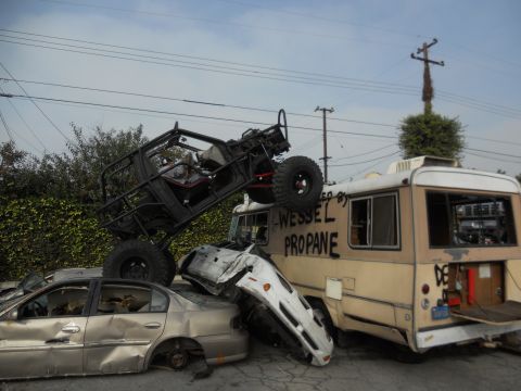 1986 Ford Bronco - An off-road vehicle is precariously balanced on a pile of wrecked vehicles, including a sedan and an RV.