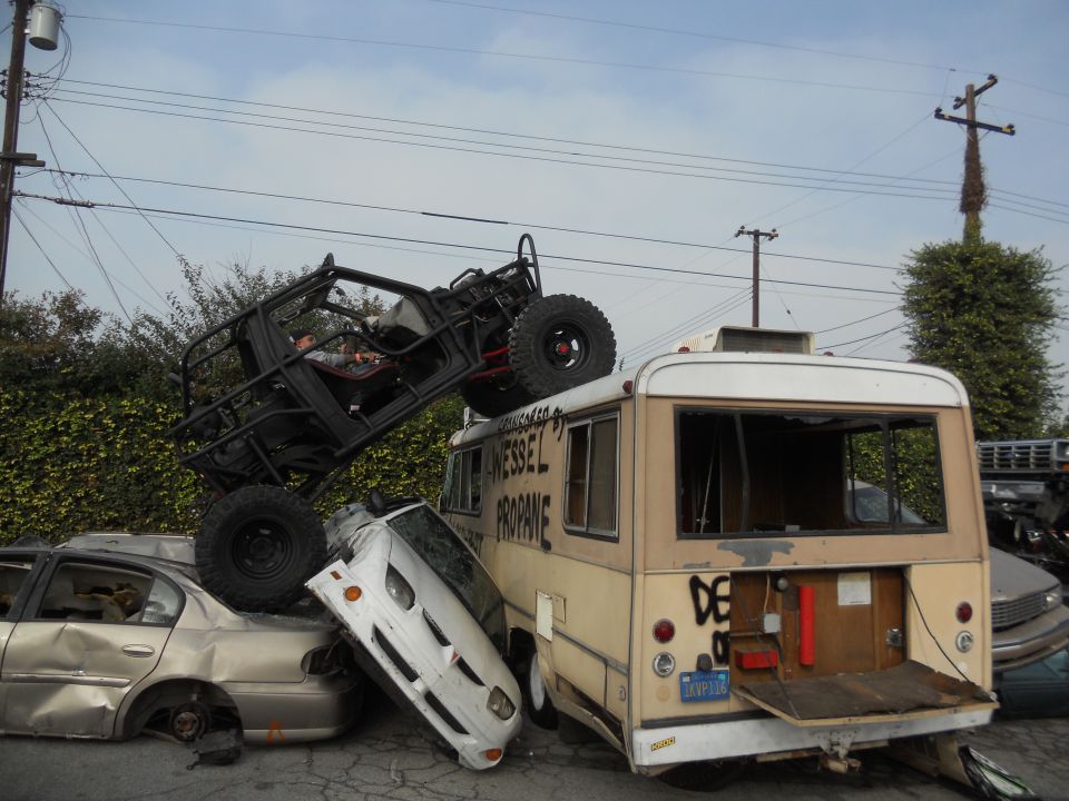 Off-road buggy on top of a wrecked bus, which is on top of other cars. Text on bus says 'WESSEL PROPANE'