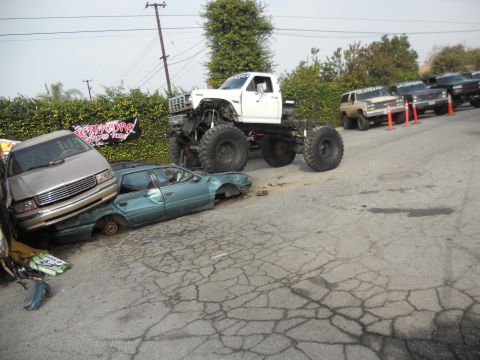1986 Ford Bronco - A monster truck looms over two crushed cars, with a line of other trucks parked in the background.