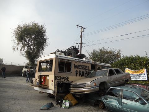 1986 Ford Bronco - Modified RV and stacked cars in a junkyard setting with graffiti and a banner.