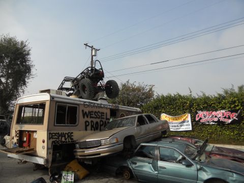 1986 Ford Bronco - A chaotic scene of stacked, damaged vehicles and a bus topped with a buggy, under a clear sky.