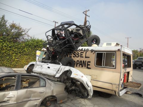 1986 Ford Bronco - A chaotic scene shows a dune buggy perched on a pile of wrecked vehicles, including an RV with 'Vessel Propane' spray-painted on its side.