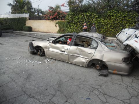 1986 Ford Bronco - A heavily damaged sedan, missing all its wheels, is seen in a parking lot next to another vehicle.