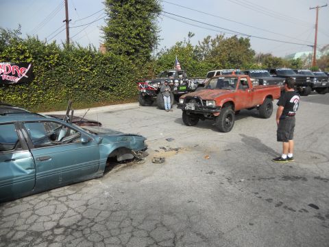 1986 Ford Bronco - A collection of modified trucks and a heavily damaged sedan are parked in a lot, suggesting a recent event or gathering.