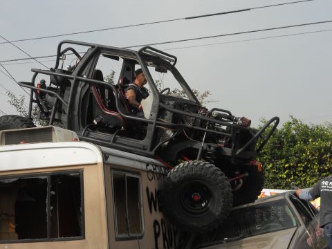 1986 Ford Bronco - A heavily modified off-road vehicle is precariously perched atop a bus, with a driver visible inside the buggy.