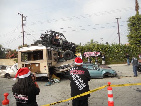 1986 Ford Bronco - A chaotic scene with multiple vehicles crushed and stacked, including a bus and a car, under a roll cage vehicle, with onlookers in festive…