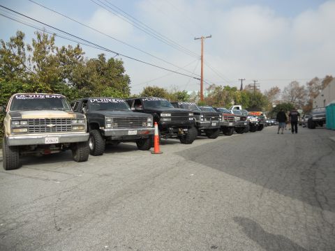 1986 Ford Bronco - A row of lifted trucks and SUVs are parked in a lot, with a 1986 pickup truck prominently featured at the front.