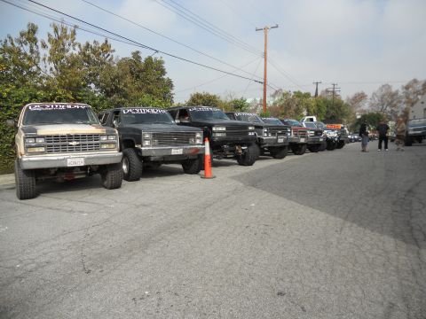 1986 Ford Bronco - A row of modified trucks and SUVs are parked on a street, some displaying banners across their windshields.